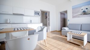 Una cocina moderna con armarios blancos, un microondas y una cafetera. Hay un comedor con una mesa y sillas a rayas. Un sofá gris y una pequeña mesa de madera están en la sala de estar. Un cuadro de una foca cuelga en la pared.