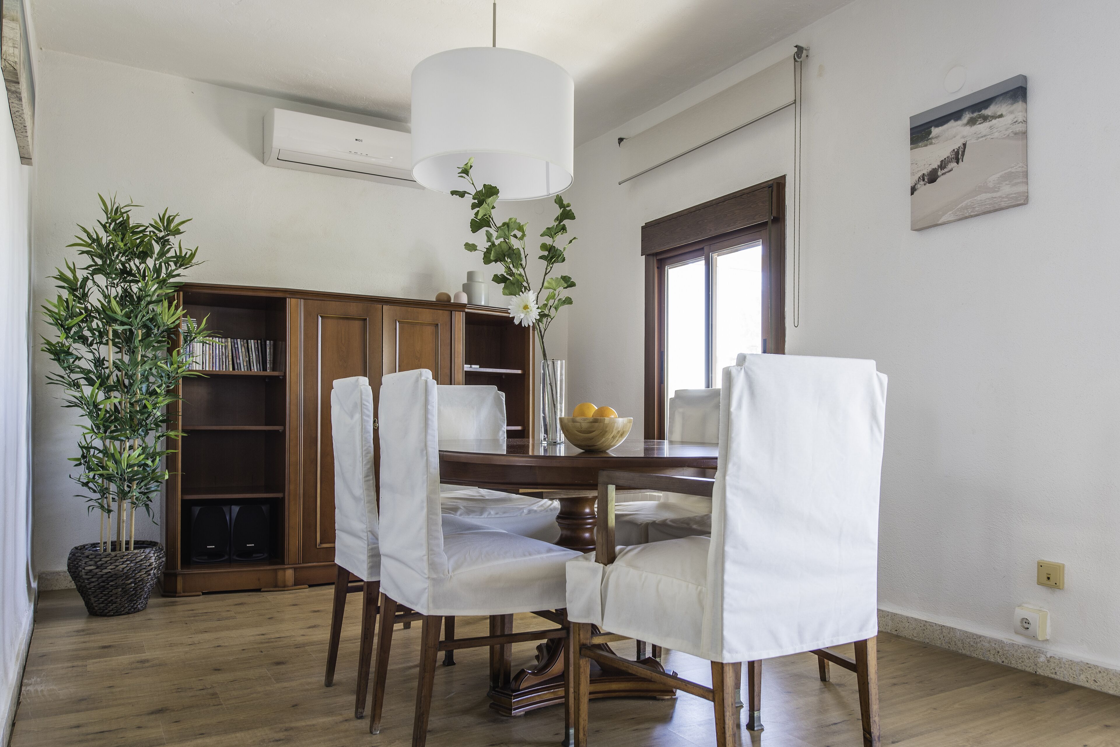A dining area with a wooden table, white chairs, a vase with flowers, and a bookshelf.
