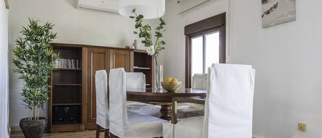 A dining area with a wooden table, white chairs, a vase with flowers, and a bookshelf.