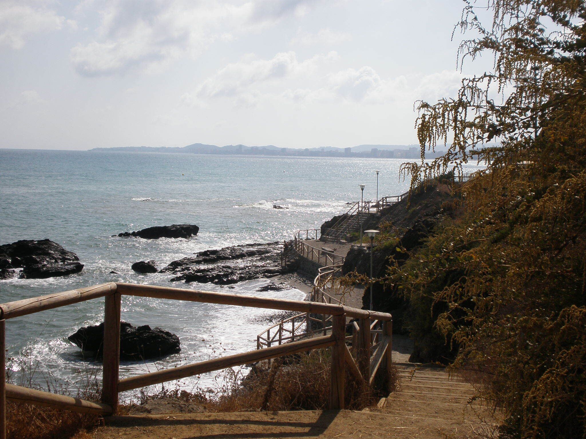 A coastal walkway with a wooden railing, rocky shoreline, and a distant city skyline.