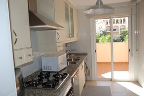 A kitchen with a gas stove, microwave, and double sink. There is a sliding glass door leading to a balcony with a view of a building and greenery.
