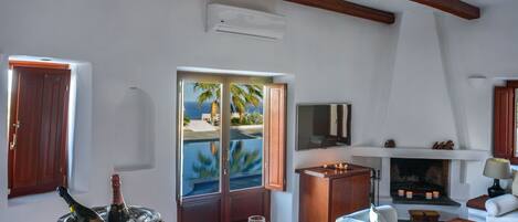 A spacious living room with a wooden ceiling, a fireplace, a television, and a view of palm trees through a door.