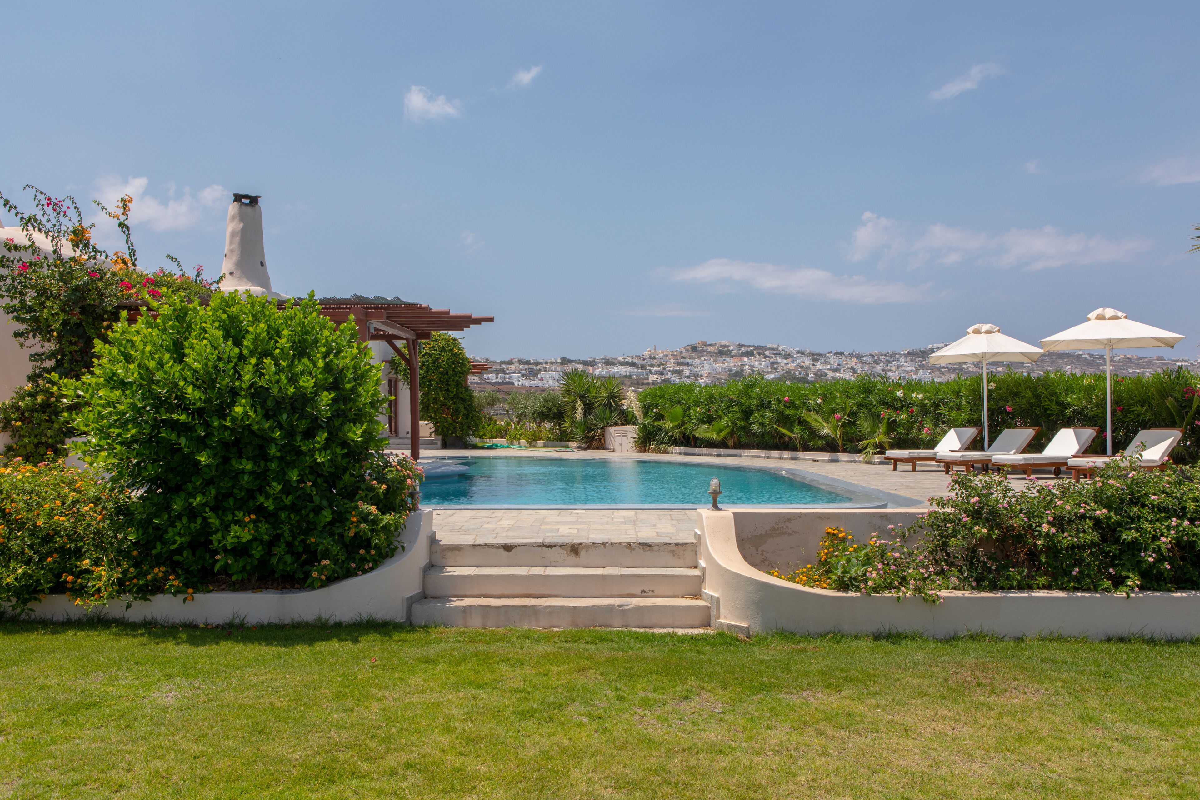 A swimming pool with steps leading down to it, surrounded by greenery and a view of a cityscape in the background.