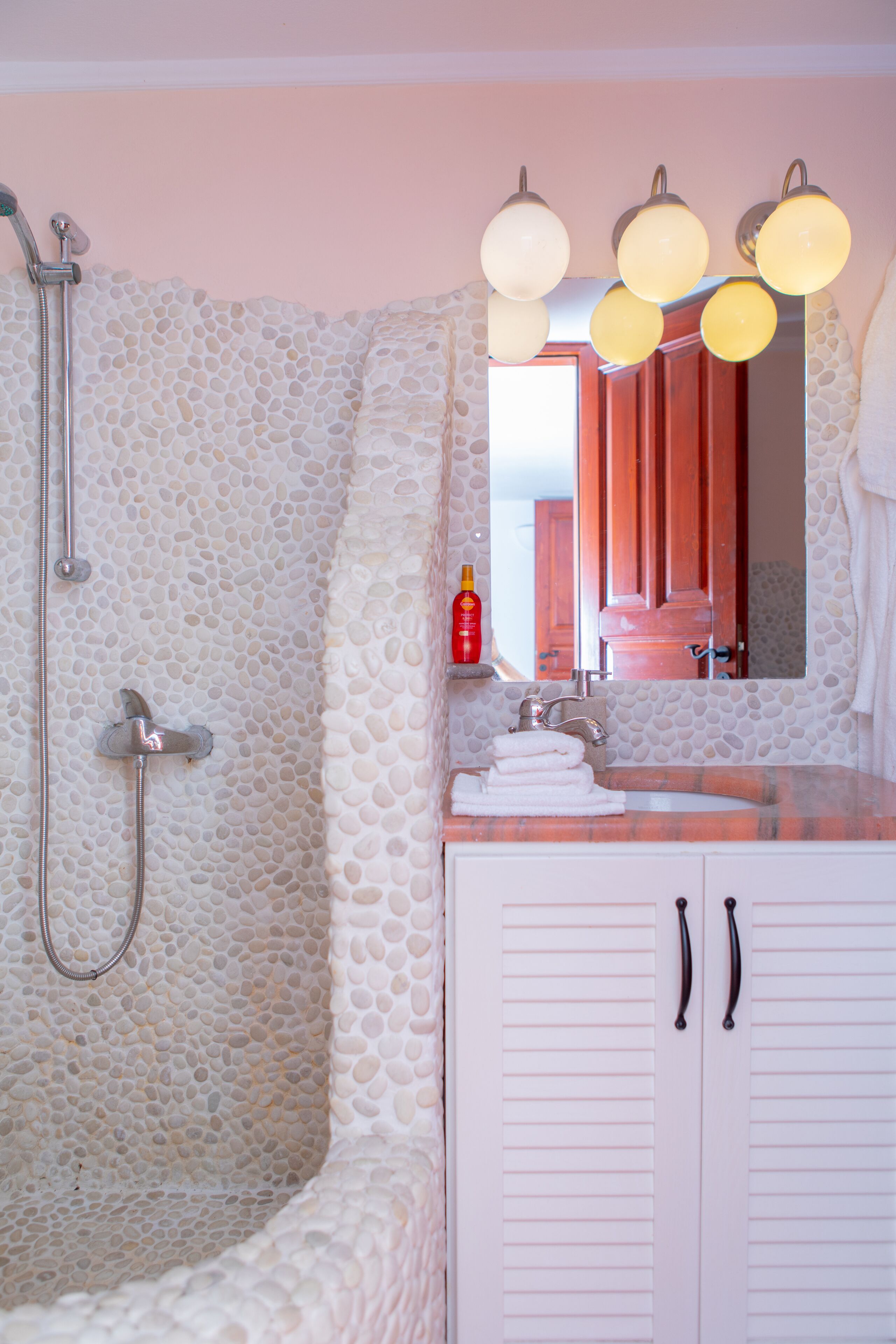 A bathroom with a stone shower enclosure, a sink with a mirror, and a red bottle on the countertop.