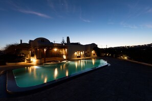 A large swimming pool with a modern design, surrounded by a paved area and a building with a dome and multiple chimneys. - Aquaria Villa (Santorini)