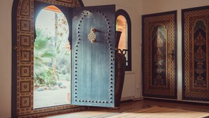 Une pièce avec une grande porte en bois, ornée de sculptures complexes, et dotée d’une poignée en métal. Un grand vase est posé sur le sol, à côté de la porte.