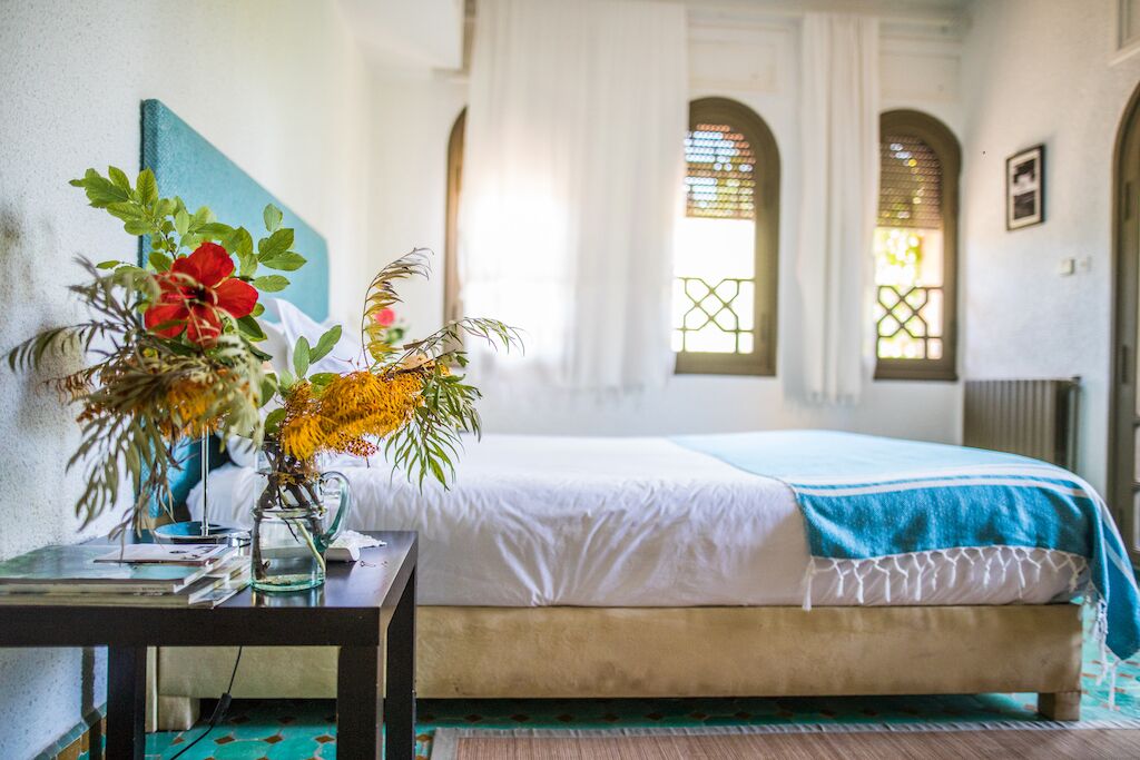 Ein Schlafzimmer mit einem Bett, einem Nachttisch mit Blumen in einer Vase und einem Fenster mit Vorhängen.