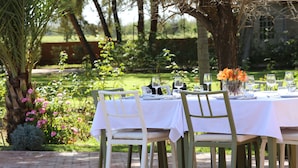 Un espace de restauration en plein air, avec des tables recouvertes de nappes blanches et des chaises vertes, entouré d’une végétation luxuriante et de plantes en fleurs.