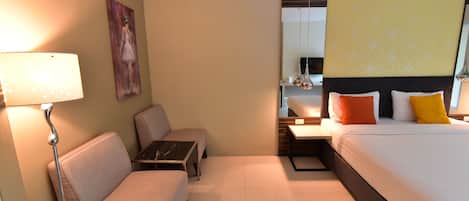 Una habitación de hotel con cama, sofá, una mesita, lámpara, un cuadro en la pared y un espejo sobre la cama.