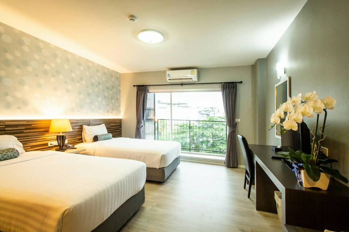 A hotel room with two beds, a desk, a chair, a lamp, and a large window with a view of the outside.