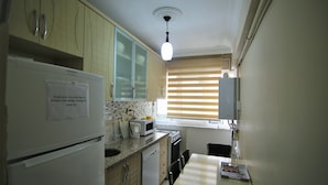 A compact kitchen with a refrigerator, microwave, and stove. There is a window with blinds, a sink, and a dining area with chairs. - Yılmaz Apart (Istanbul)