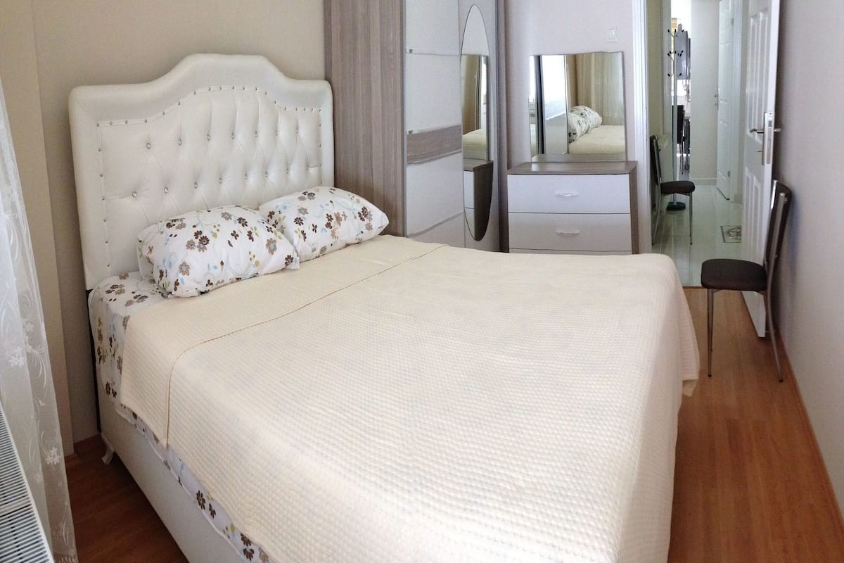 A bedroom with a tufted headboard, floral bedding, a bedside table, a mirror, and a chair.