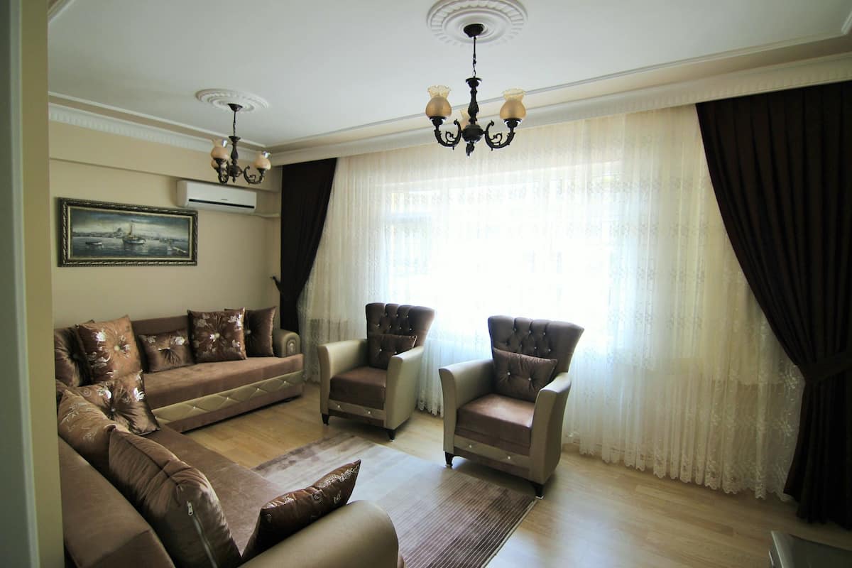 A living room with a brown sofa set, two armchairs, a chandelier, and a large window with curtains.