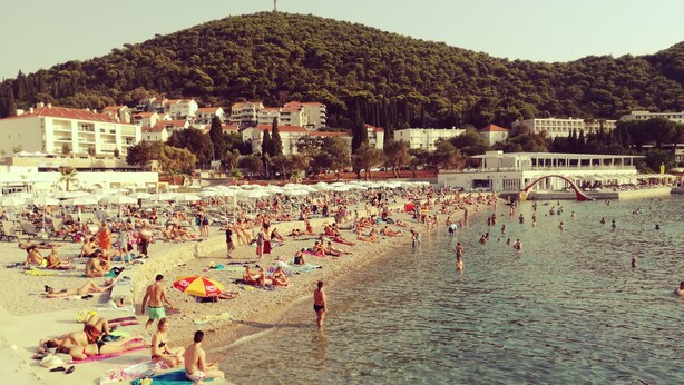 Ein überfüllter Strand mit Menschen, die sich sonnen und schwimmen, umgeben von Gebäuden und einer Hügelkette im Hintergrund.