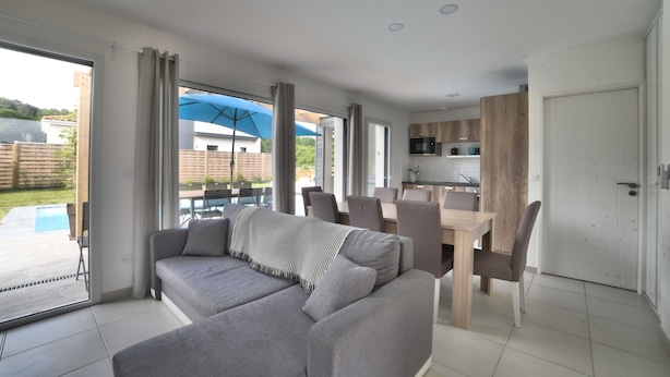 A modern living room with a grey sofa, dining table, and chairs. A kitchen area with wooden cabinets and a microwave is visible. - Villa 4 by Avecoeur (Saint-Augustin)
