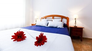 Un letto rifatto con una coperta blu, cuscini bianchi e due fiori rossi. Sono presenti due comodini con lampade, una testiera in legno e una parete bianca con un punto luce.