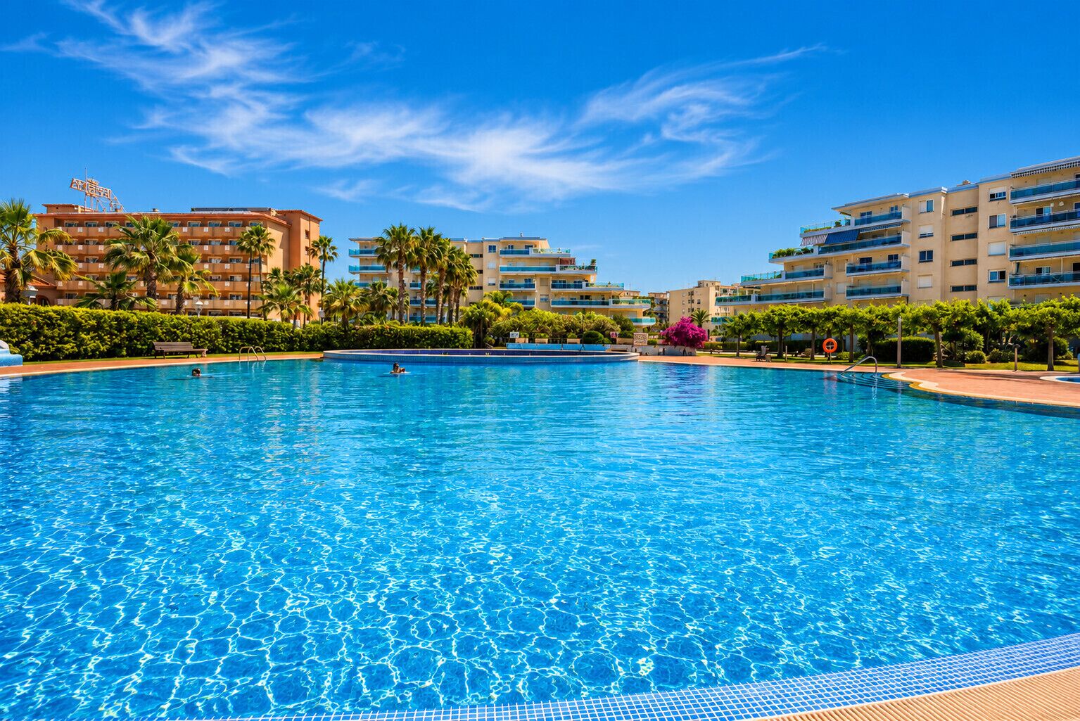 A large swimming pool surrounded by multi-story buildings with balconies, palm trees, and a clear blue sky.