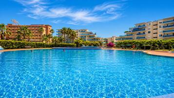 A large swimming pool surrounded by multi-story buildings with balconies, palm trees, and a clear blue sky.