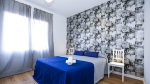 Un dormitorio con una cama, una silla, una mesa con una toalla, un ventanal con cortinas y una pared con diseño.