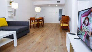 Una sala de estar moderna con un sofá gris, una mesa de comedor con sillas, un televisor sobre un mueble blanco y suelo de madera.