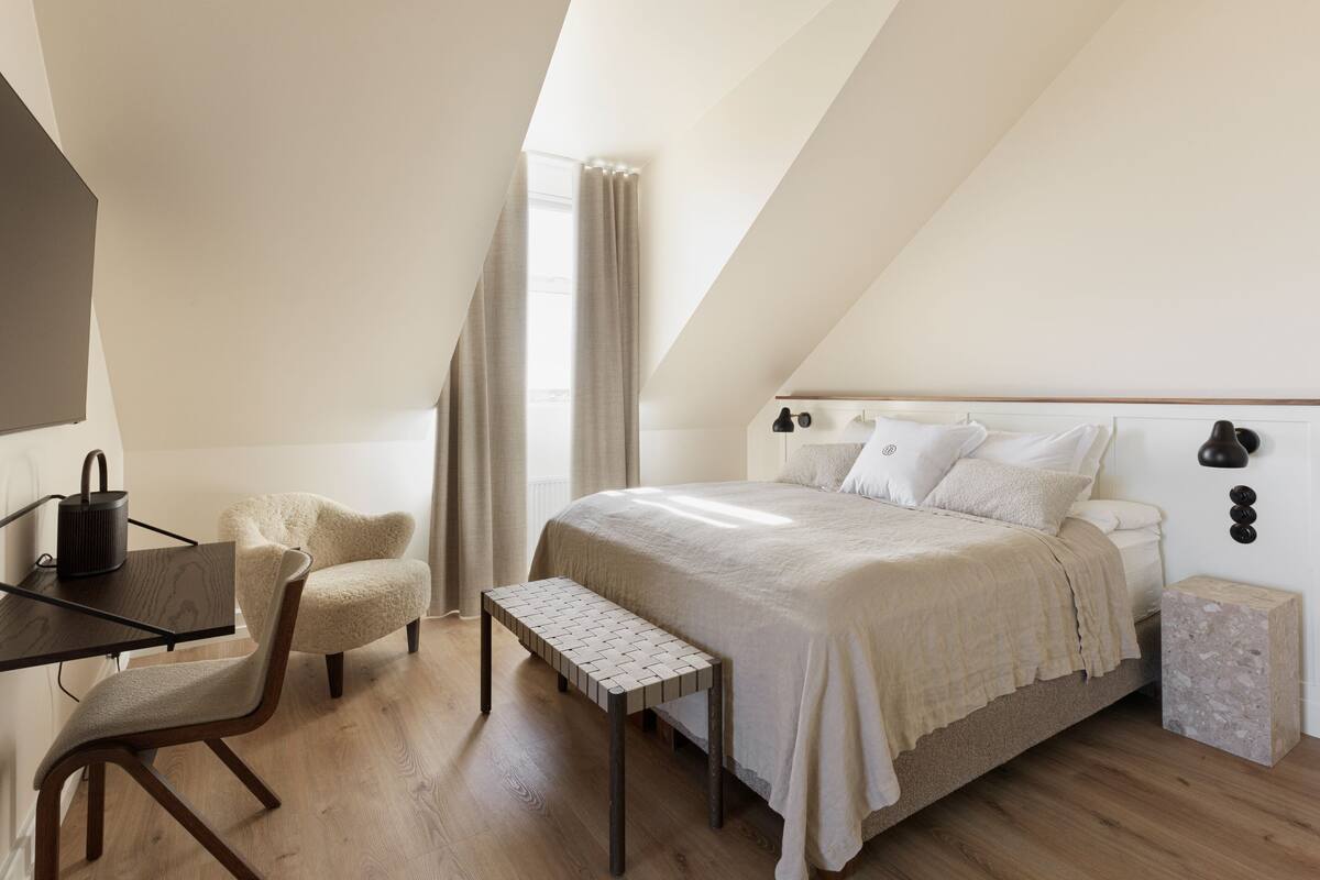 Un dormitorio con techo inclinado, una cama con cabecera, un escritorio con silla, una ventana con cortinas y una mesita de noche pequeña.