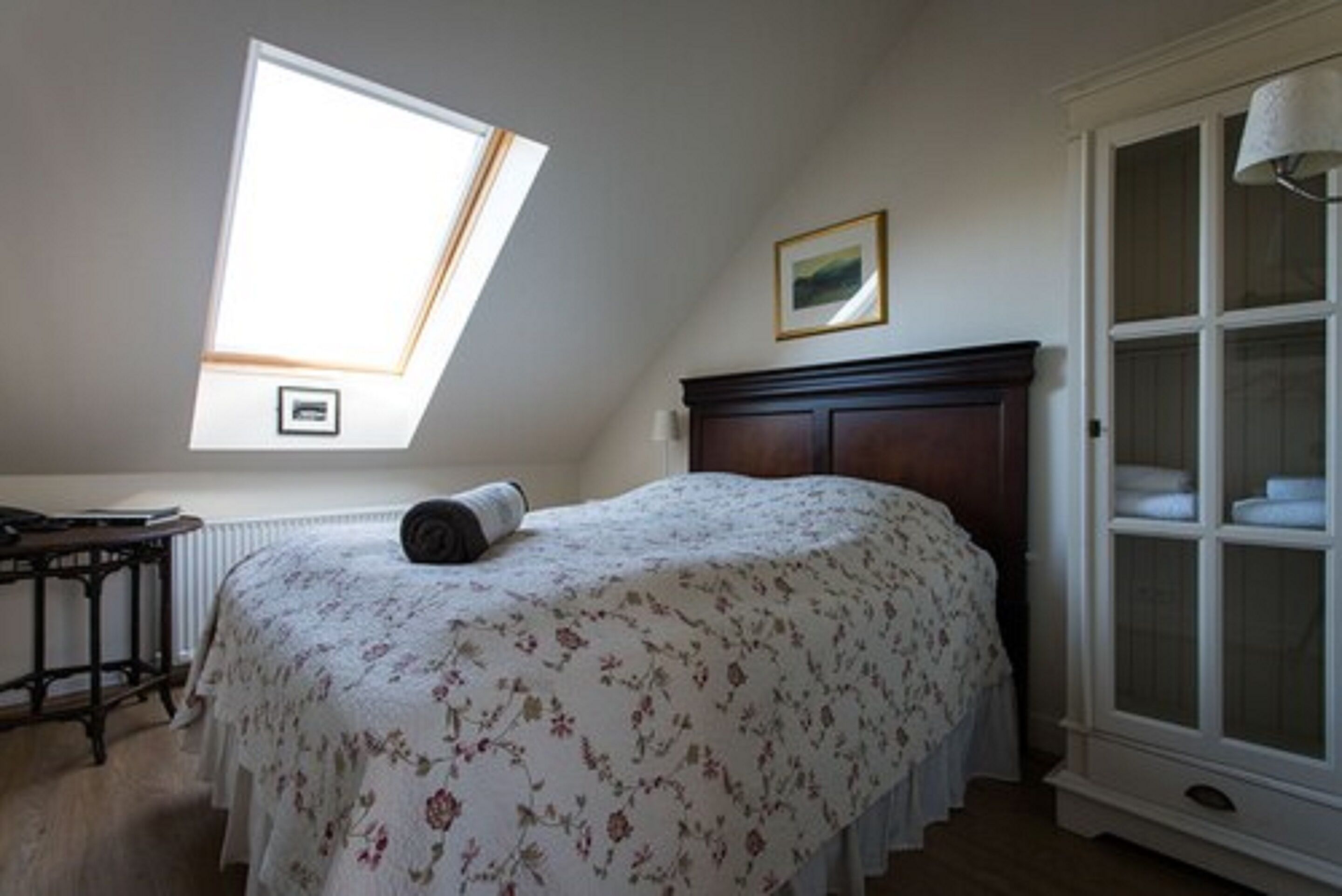 A bedroom with a sloped ceiling, a bed with a floral bedspread, a wooden nightstand, a dresser with glass doors, and a framed picture on the wall.