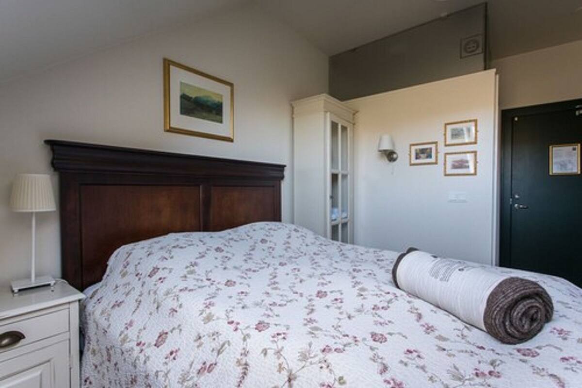 Un dormitorio con cama, mesita de noche, lámpara, puerta y una pared con cuadros.