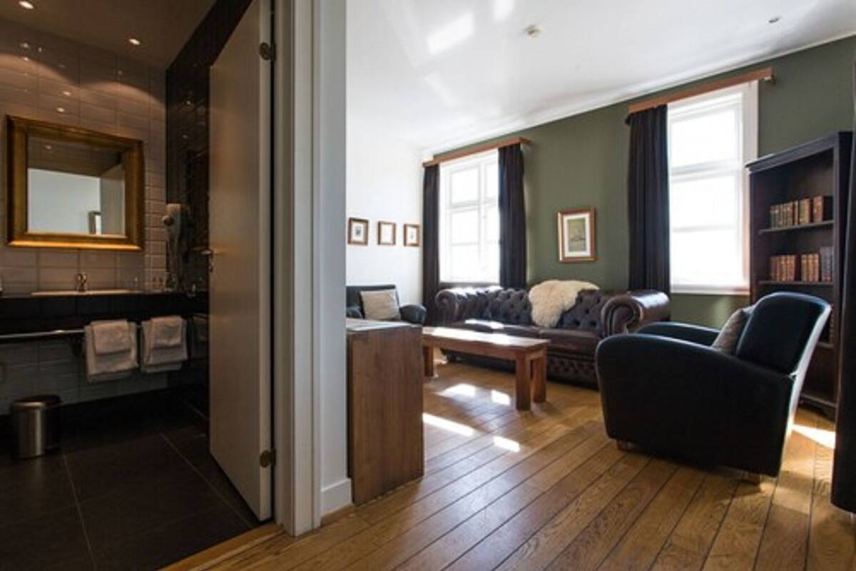 Una sala moderna con sofá, sillón y una mesa de centro. Se observa una estantería y una ventana con cortinas.