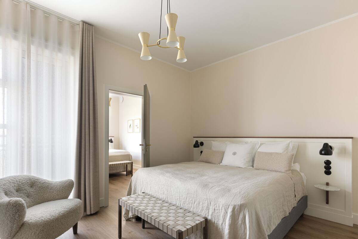 Un dormitorio con cama, una silla, un banco, una mesita de noche y una lámpara colgante.
