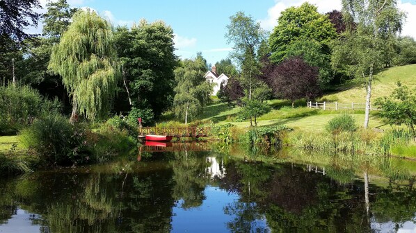 A serene lakeside setting with a wooden bridge, a small red boat, and a house on a hill. - Charming English cottage, Ashdown Forest, excellent pub (Danehill)