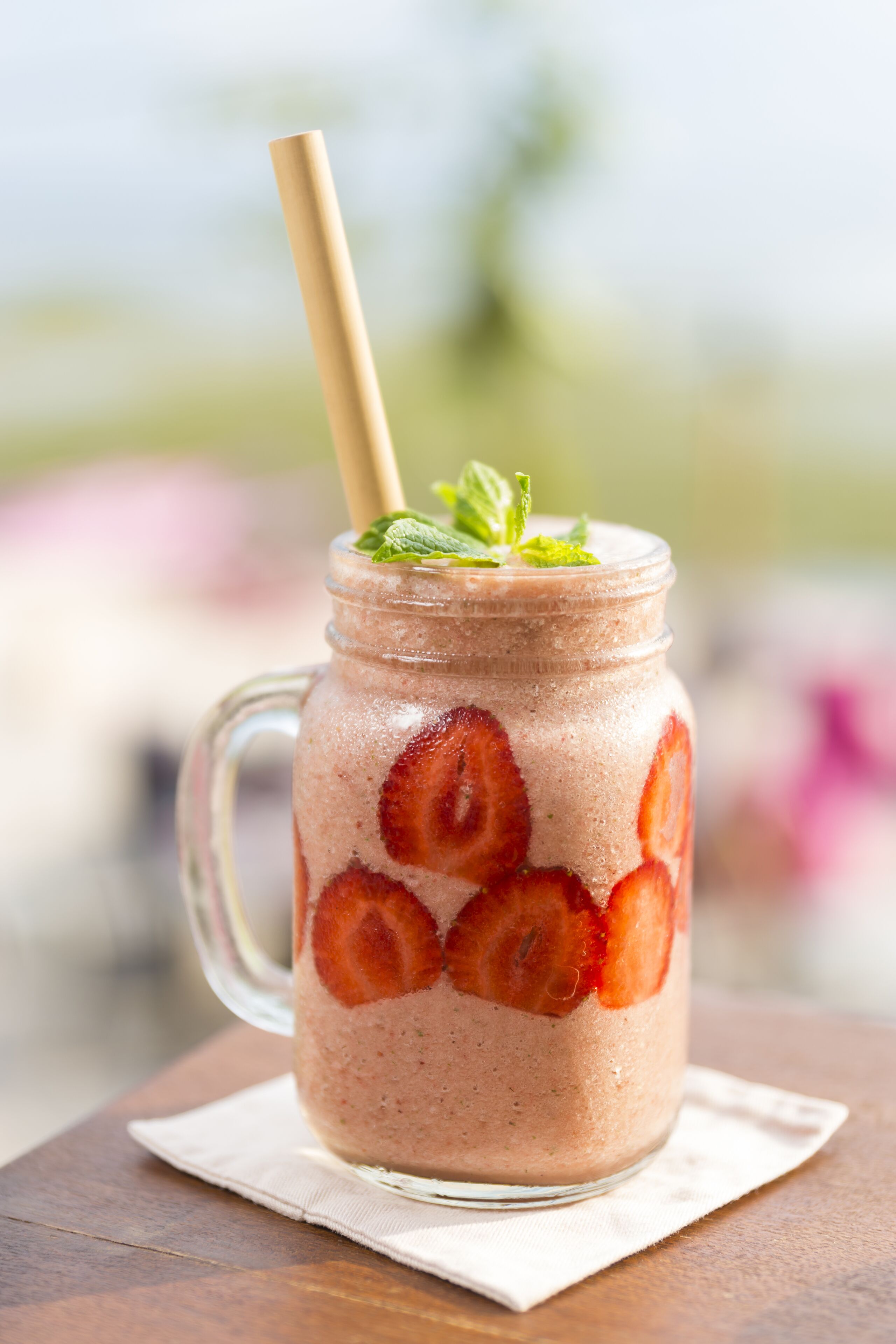 A glass mug with a straw, filled with a pink beverage and garnished with strawberries and mint leaves.