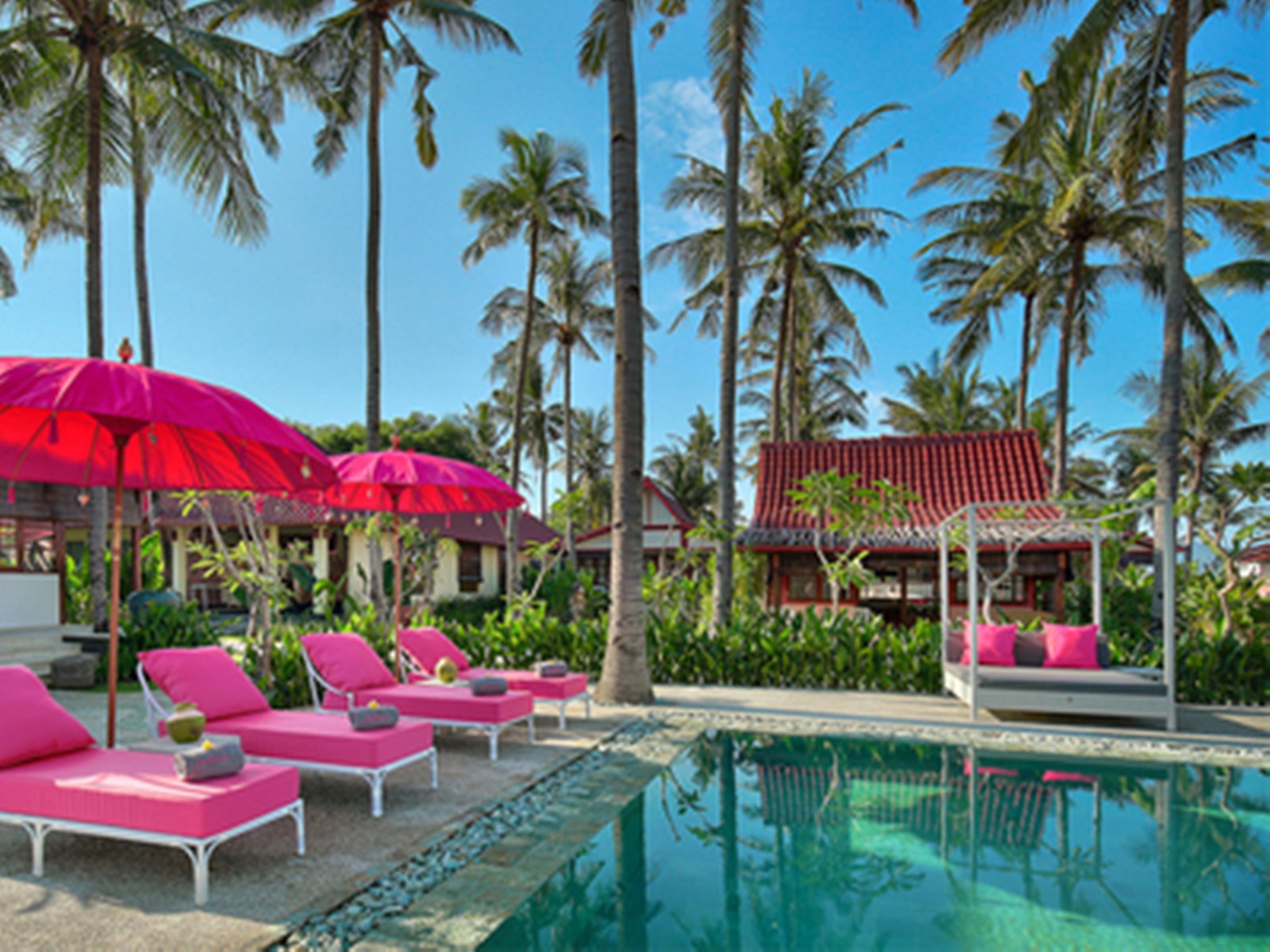 A poolside area with lounge chairs and umbrellas, surrounded by palm trees and a building with a red roof.