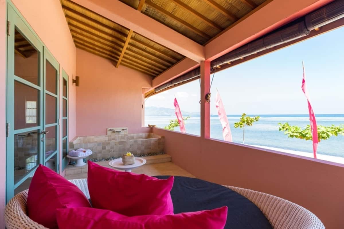 A balcony with a view of the sea, a wicker chair with pillows, a small table with a cake, and a bathtub.