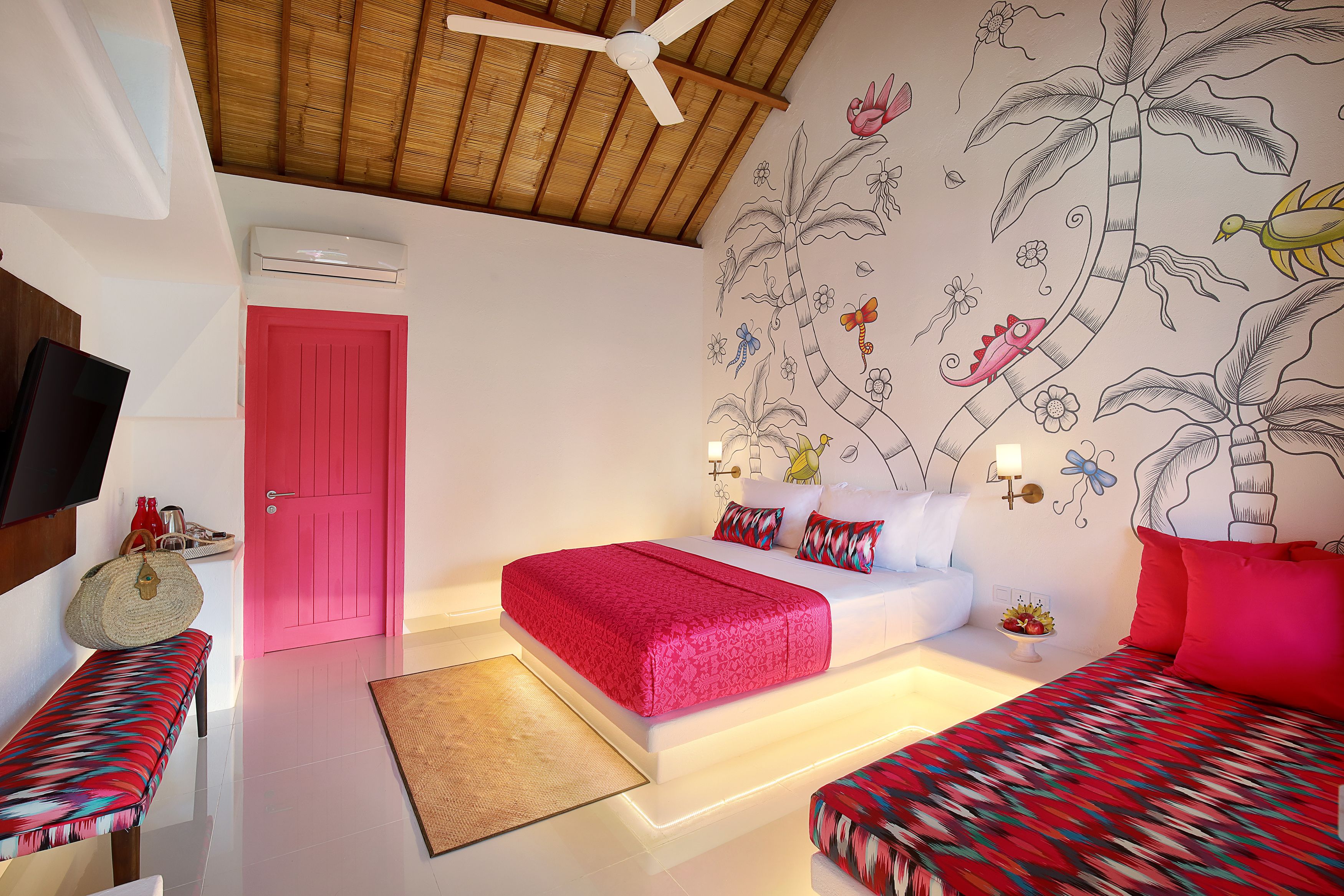 A hotel room with a wooden ceiling, a pink door, a bed with red bedding, a bench, and a wall mural.