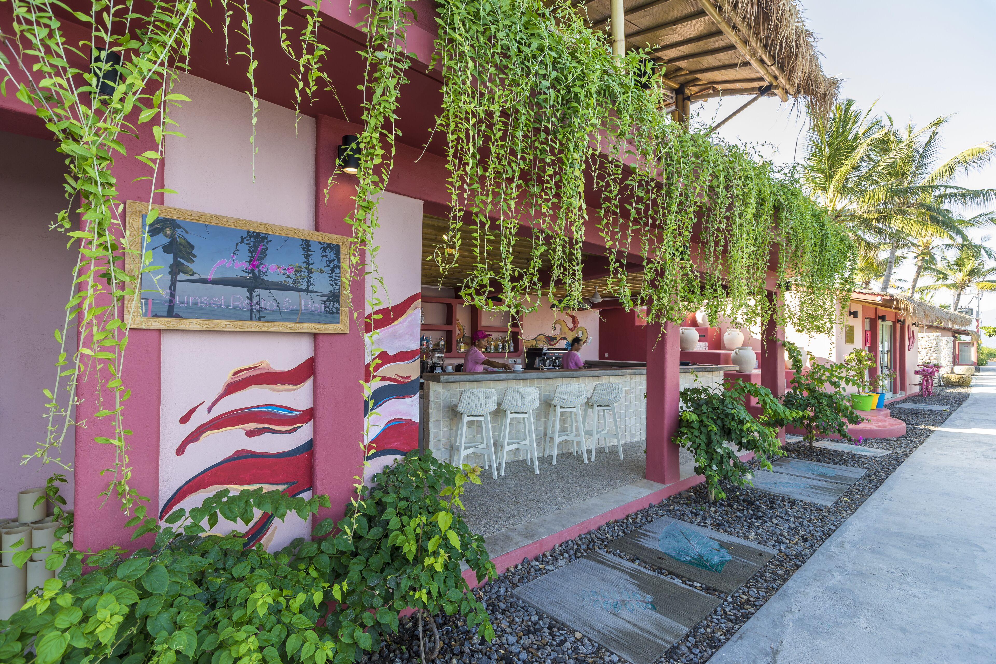 A bar area with white bar stools, a pink wall with a mural, and hanging greenery.
