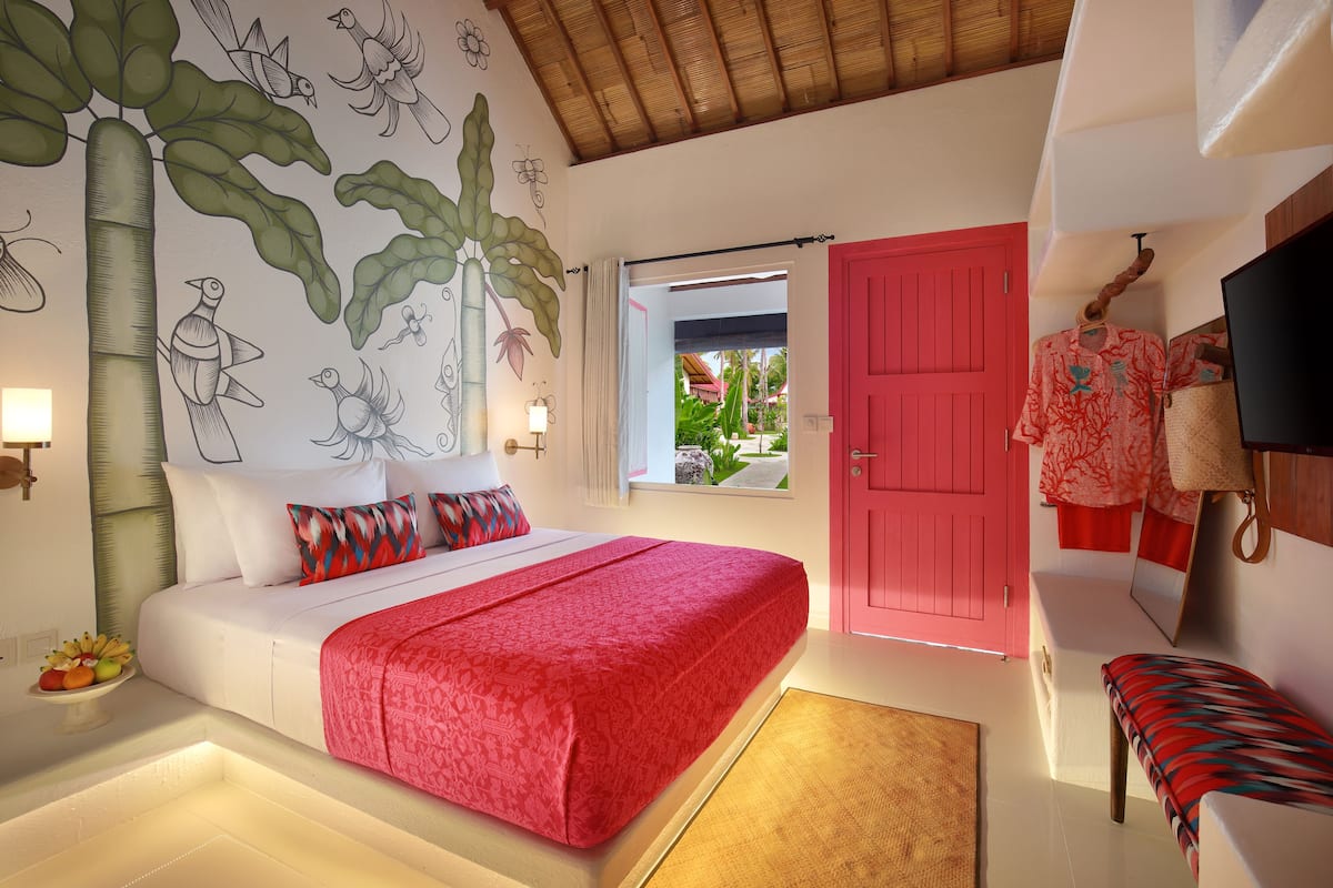 A bedroom with a bed, a red door, a window, and a wall mural.