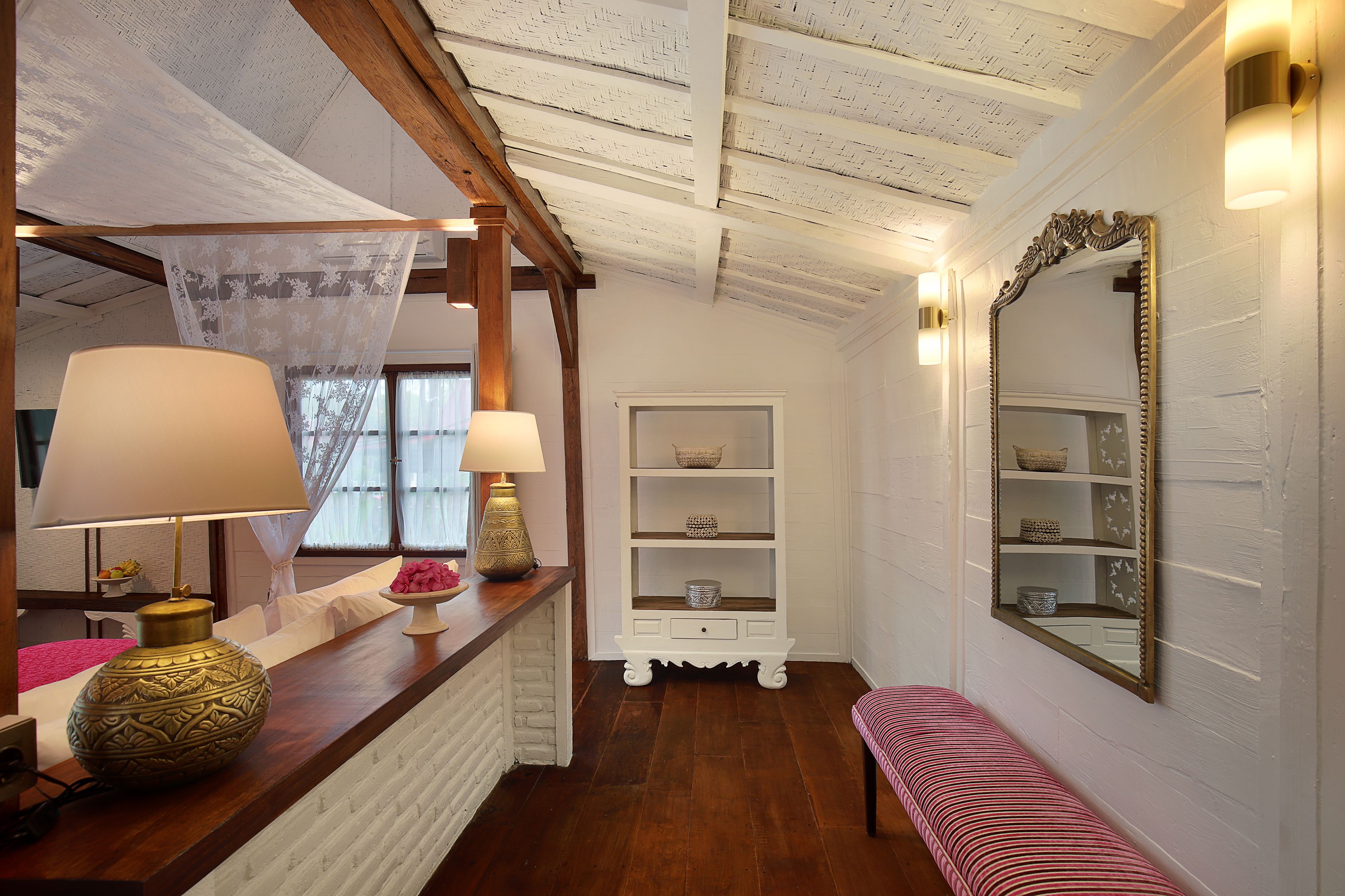 A room with a wooden ceiling, a white wall, a bench, a mirror, and a shelf with decorative items.