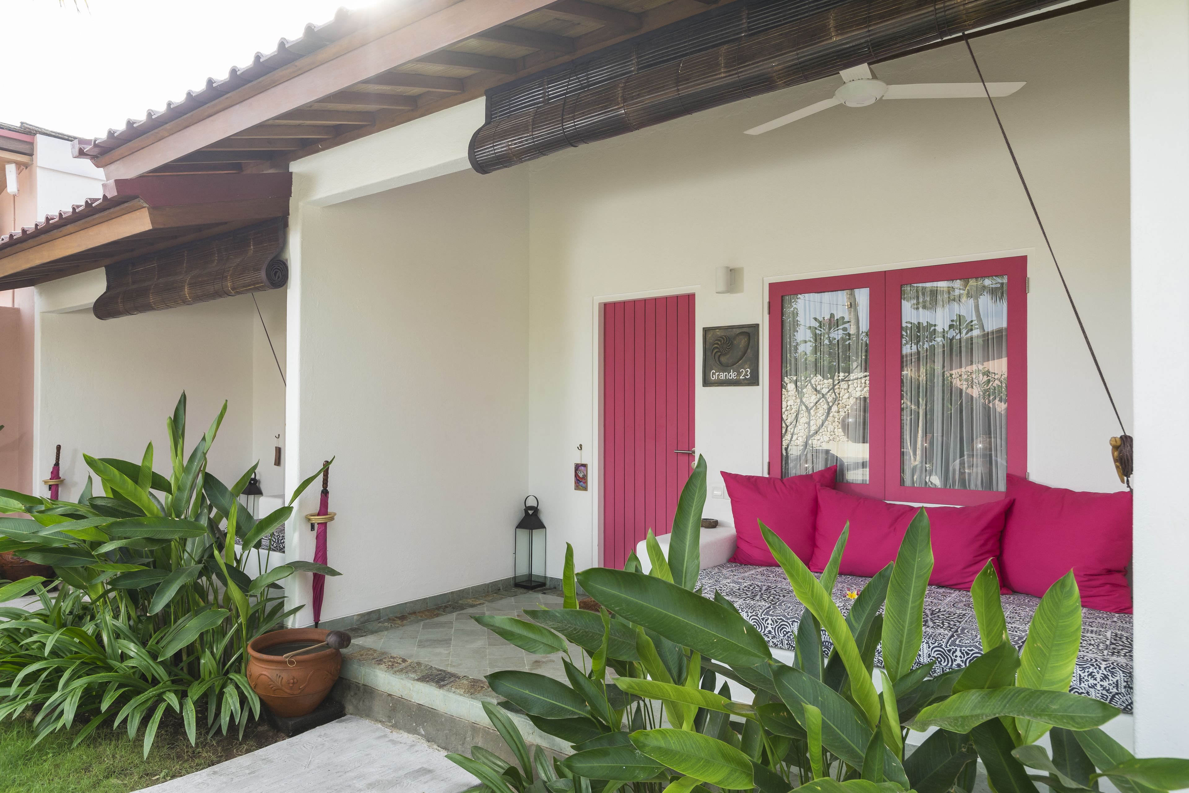 Un espace extérieur accueillant, avec une porte rouge, un ventilateur de plafond et de nombreuses plantes vertes.