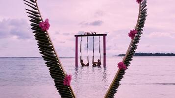 A heart-shaped structure made of palm leaves and pink flowers, with a swing set in the center, overlooking a beach.