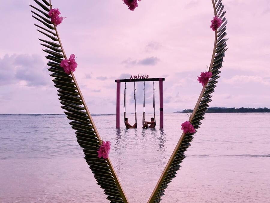 A heart-shaped structure made of palm leaves and pink flowers, with a swing set in the center, overlooking a beach.