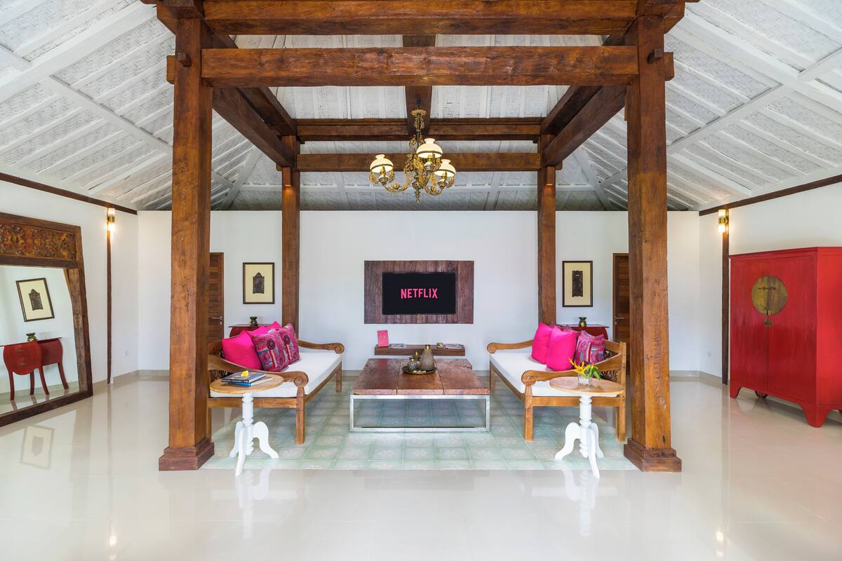 A spacious lobby with a high ceiling, wooden beams, a central coffee table, two sofas with cushions, a red cabinet, and a TV screen displaying "NETFLIX".