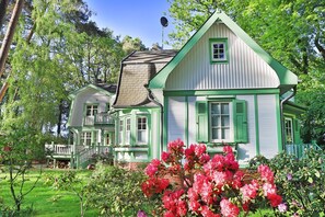 Una casa de dos pisos con adornos verdes y techo de ladrillo, rodeada de abundante vegetación y plantas florecientes.