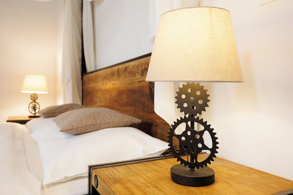 Een bed met een houten hoofdbord, een nachtkastje met een lamp met tandwielen, en een nachtkastje met een lamp.