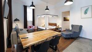 A hotel room with a wooden dining table, two chairs, a sofa, a bed with a canopy, a chair, a lamp, and a framed picture on the wall.