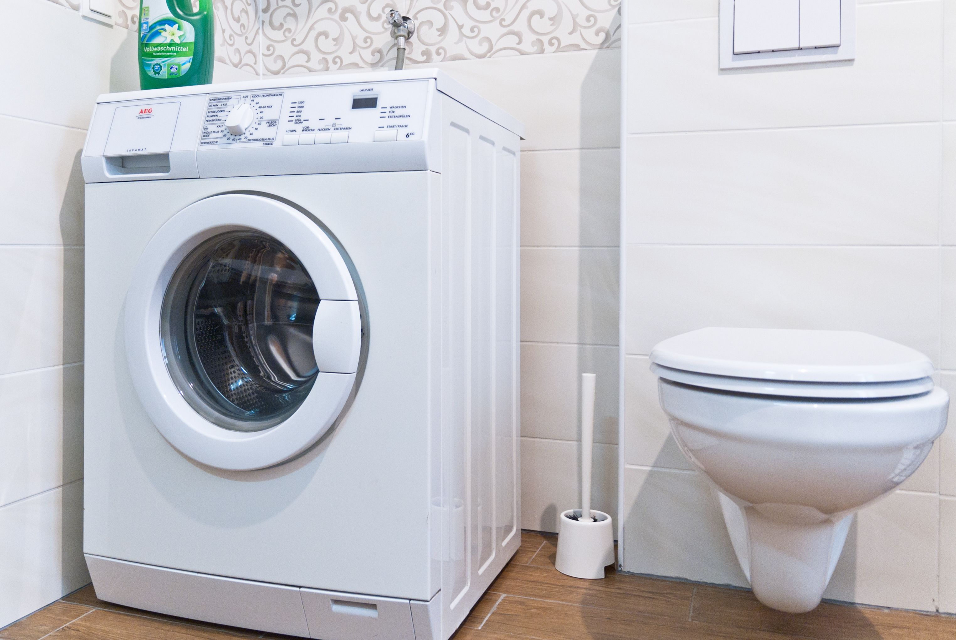 Une machine à laver à chargement frontal, placée à côté des toilettes dans une salle de bain dont le sol est recouvert de carreaux décoratifs.