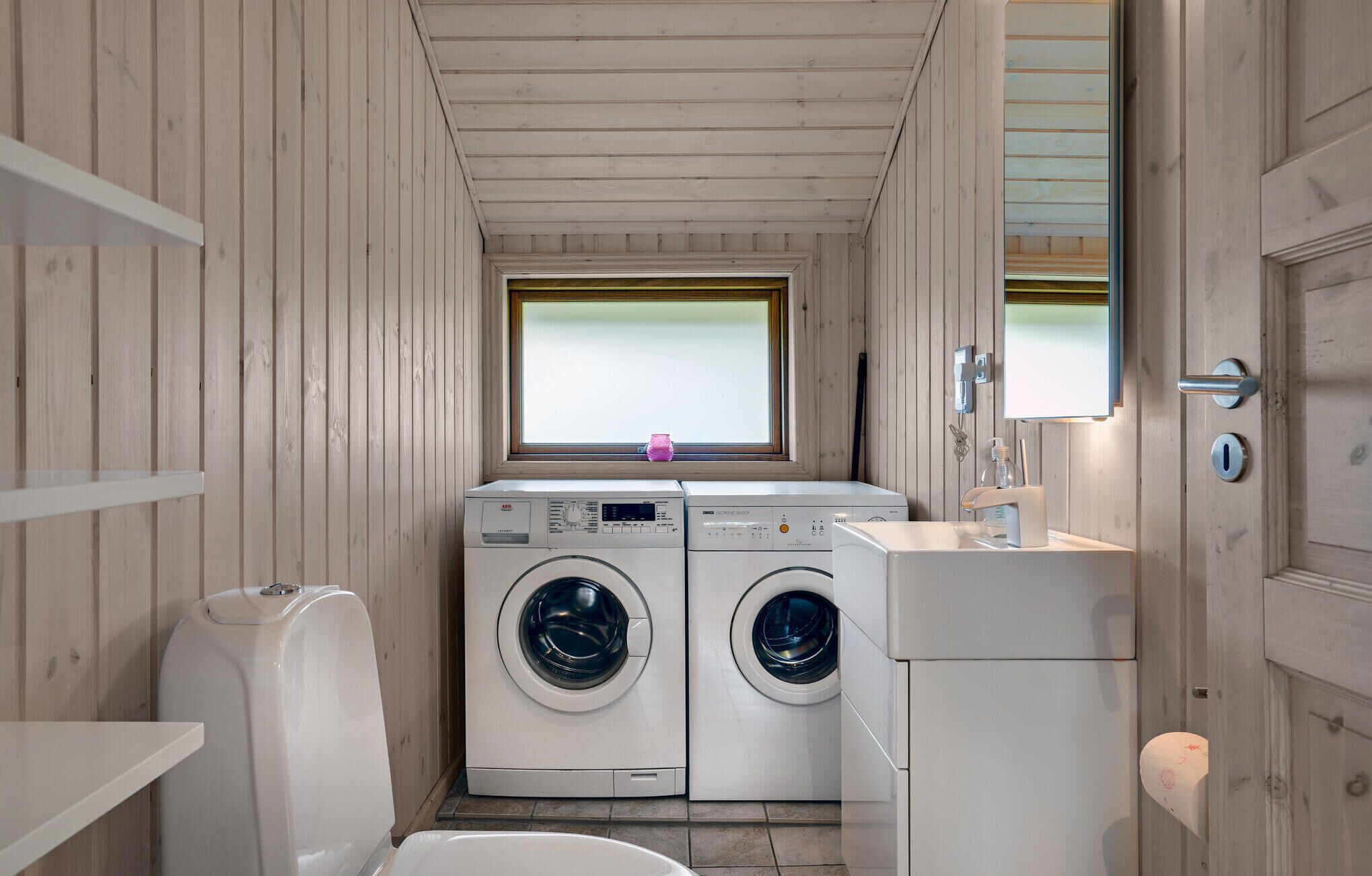 A compact laundry room with two front-loading washing machines, a sink, and a window above the machines.