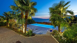 Piscina com vista para o mar, rodeada por palmeiras e um caminho pavimentado.