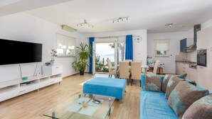 Uma sala de estar moderna com uma televisão de tela plana, uma mesa de centro de vidro, um sofá e uma área de jantar com mesa e cadeiras.