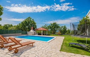 Una piscina con sillas de descanso, un pabellón y un trampolín en un jardín.