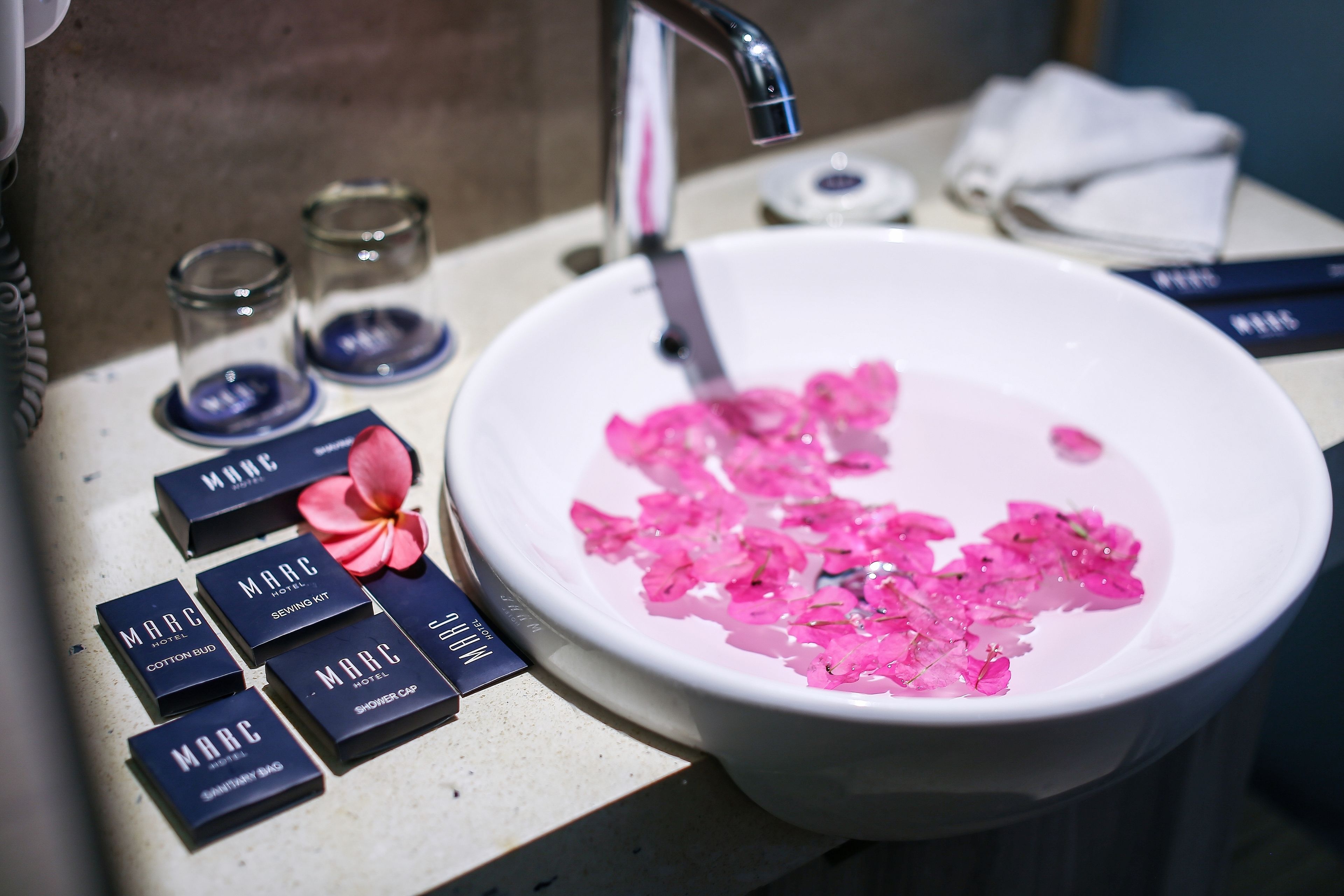 En handfat i badrummet med rosa blomblad, en handduk och flera produkter med varumärket Marc Hotel.